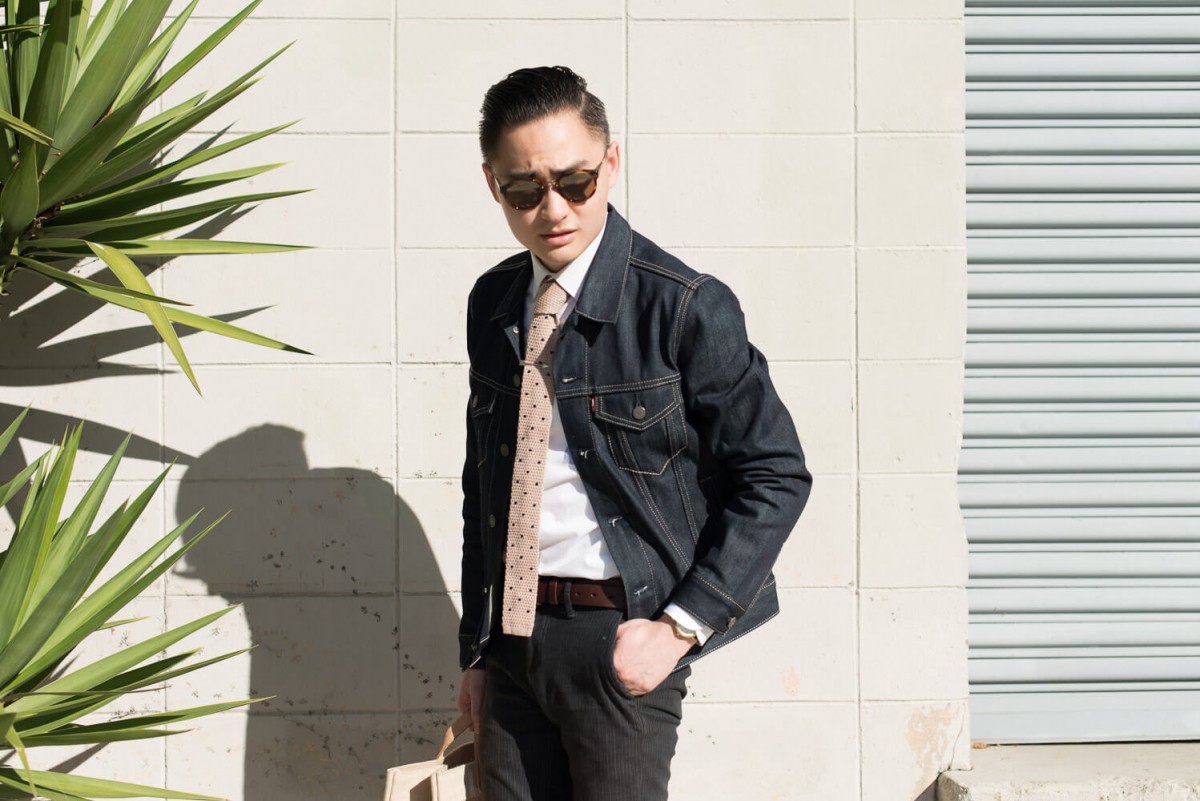 Seorang pria mengenakan jaket jeans hitam, kemeja putih, dan dasi bermotif.